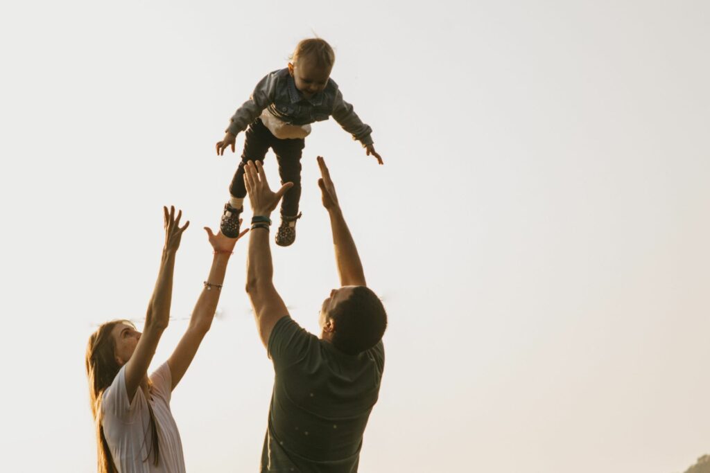 Ein herzerwärmender Moment einer Familie im Freien: Ein Kind wird lächelnd von einem Elternteil hoch in die Luft geworfen, während der andere Elternteil die Arme ausstreckt, um es aufzufangen. Der Hintergrund ist minimalistisch mit einem hellen, natürlichen Licht, das die Szene umrahmt und den Fokus auf die Freude und den Zusammenhalt der Familie legt.