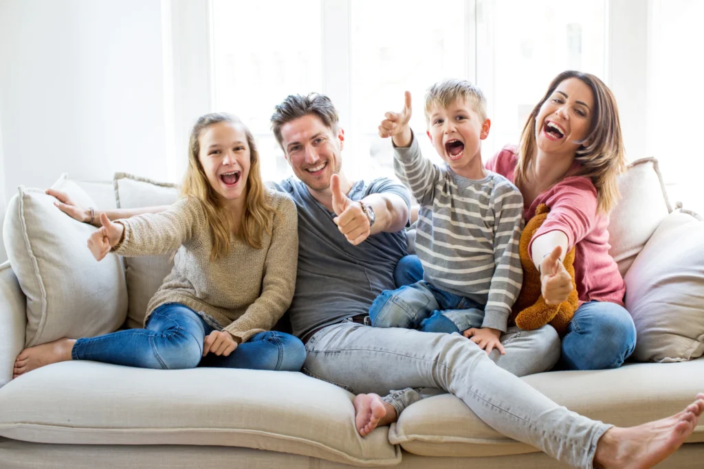 Eine glückliche Familie sitzt zusammen auf einem Sofa. Mutter, Vater, Tochter und Sohn lächeln fröhlich in die Kamera und zeigen Daumen hoch. Die entspannte Atmosphäre und das helle Tageslicht im Raum vermitteln Freude und Zusammenhalt in der Familie.